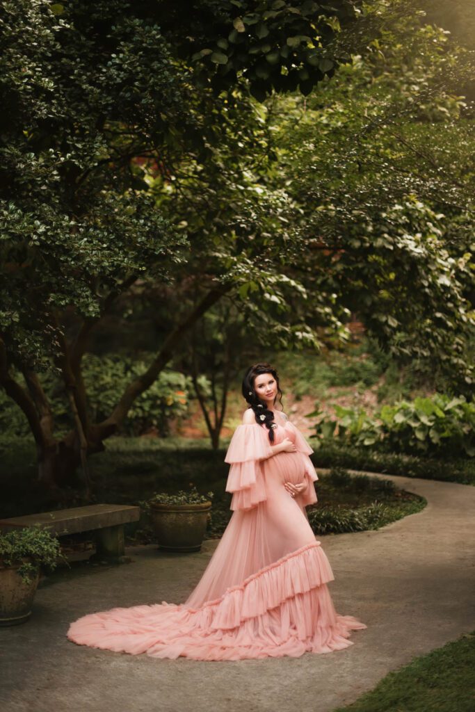 Expecting mother in a flowing blush gown standing on a curved garden path beneath dense green trees.