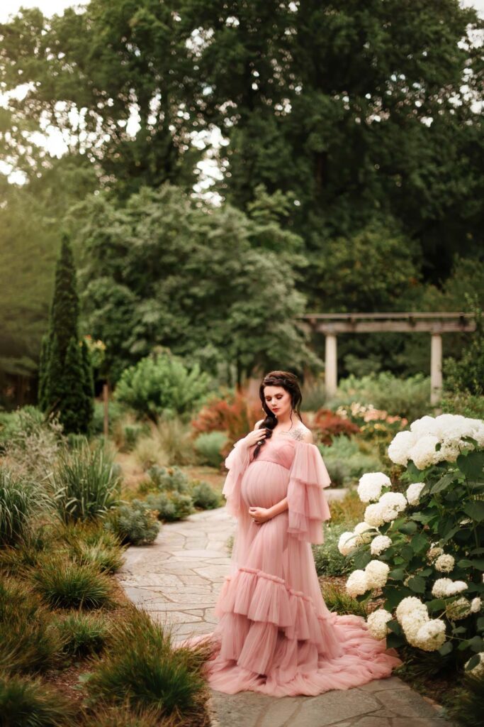 Mother cradling her belly in a blush off-the-shoulder gown in front of Pavilion beside white hydrangeas.