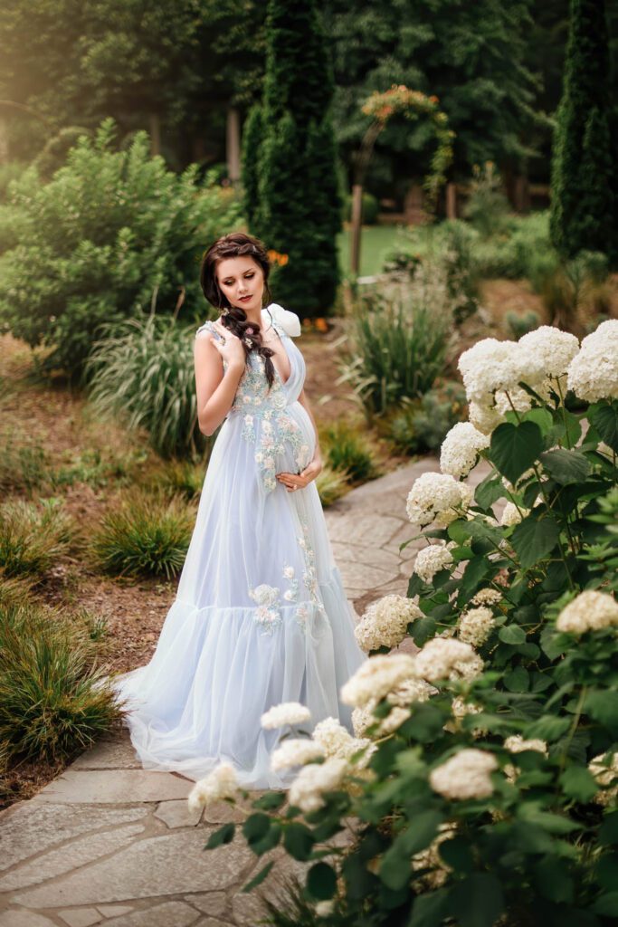 pregnant mom in a pale blue floral gown beside white hydrangeas with the Cator Woolford Gardens pavilion in the background in Atlanta, GA