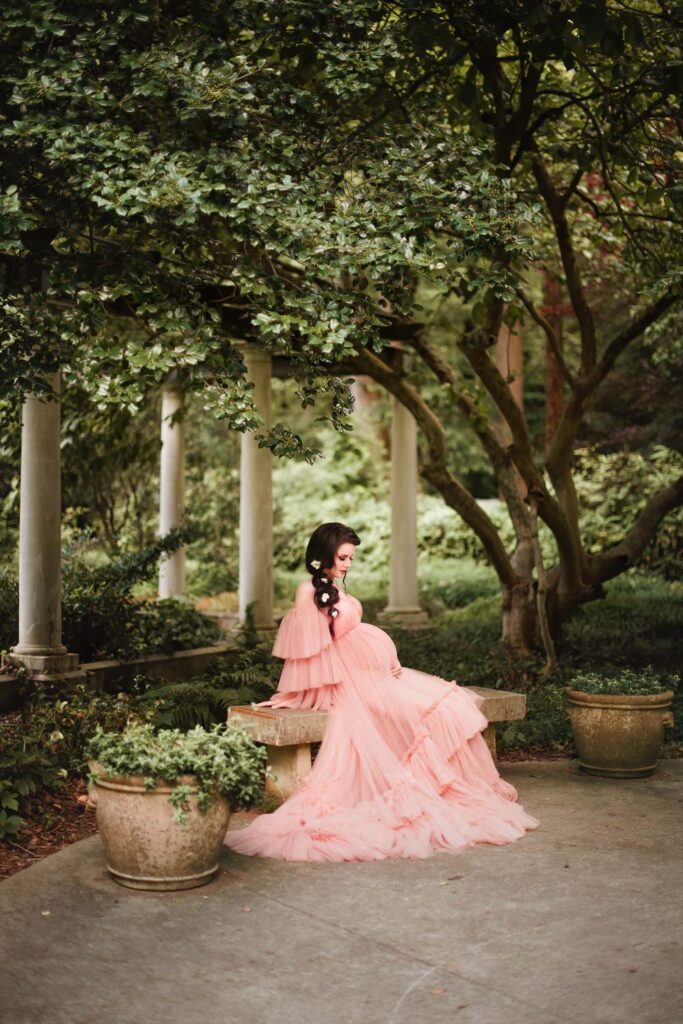 Expecting mother in blush ruffled gown seated on a stone bench with stone colonnade in the background.