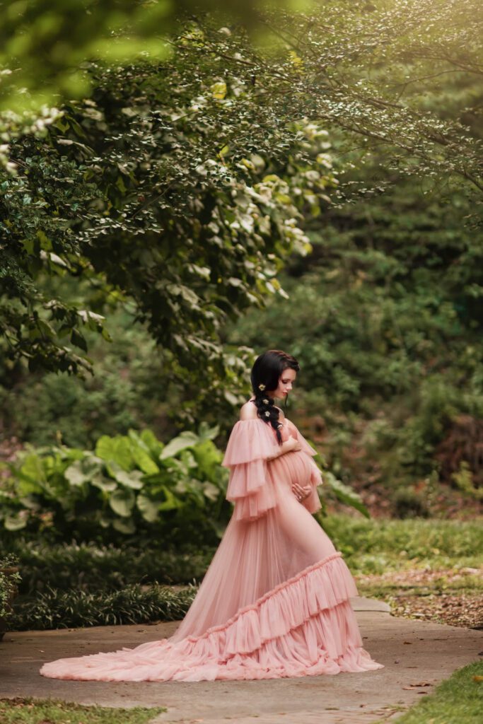 expecting mom in a flowing blush ruffled gown cradling her belly beneath arching green branches