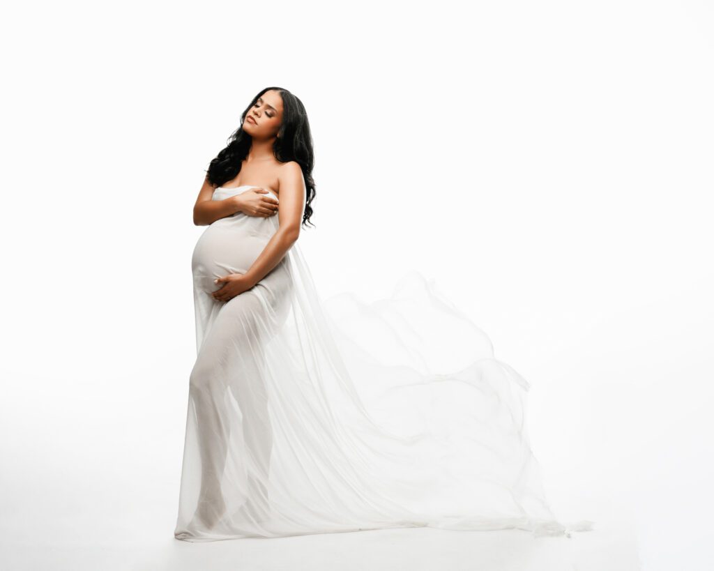 Expecting mother in a dreamy flowing white gown cradling her belly against a bright white studio background.