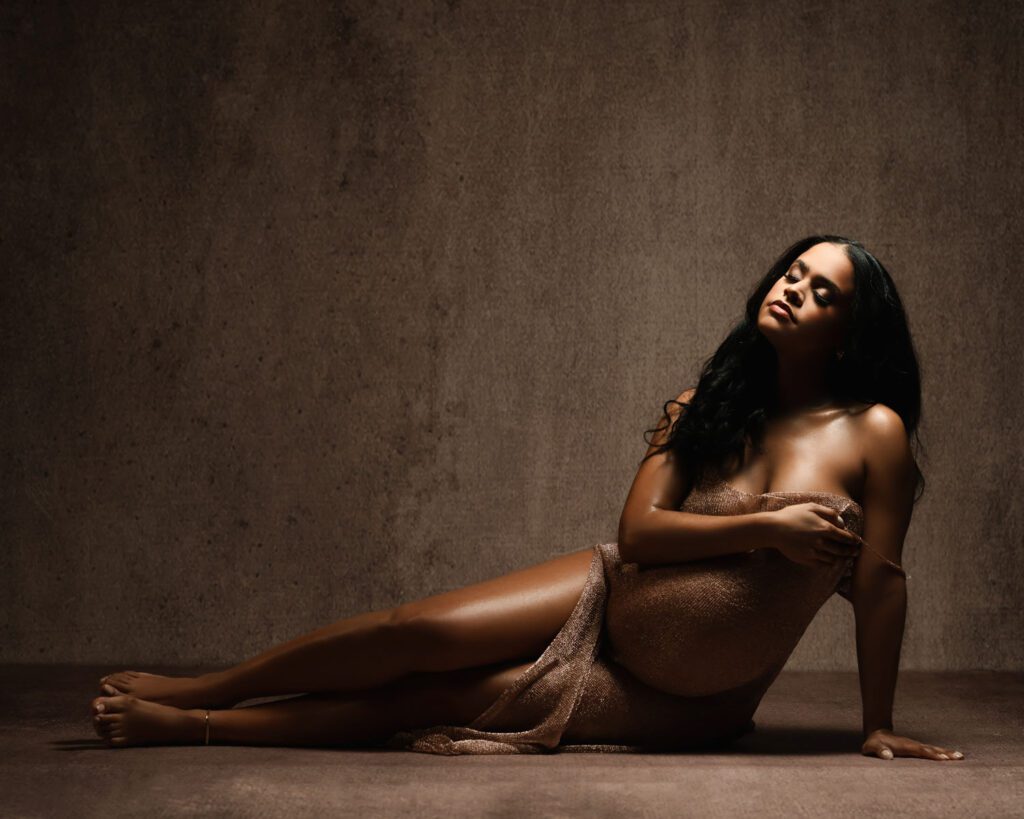 Pregnant black woman in fitted sheer dress with one strap off shoulder on laying on ground propped up with arm.