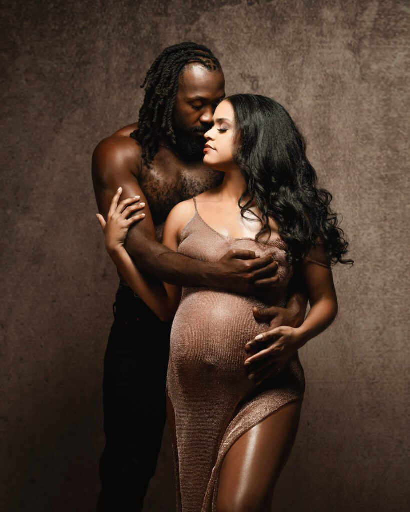 Expecting african american mother in a bronze gown embraced by a shirtless husband while both cradle her belly against a brown backdrop.