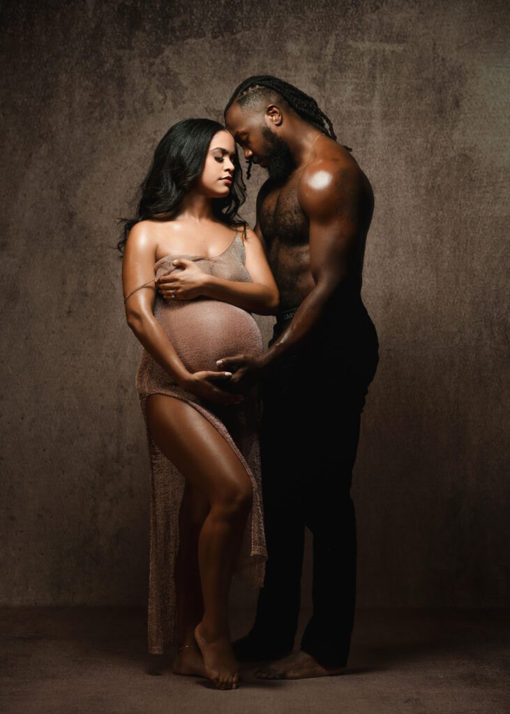 Pregnant black woman in a fitted bronze gown standing close to shirtless husband during a Marietta maternity photography session.