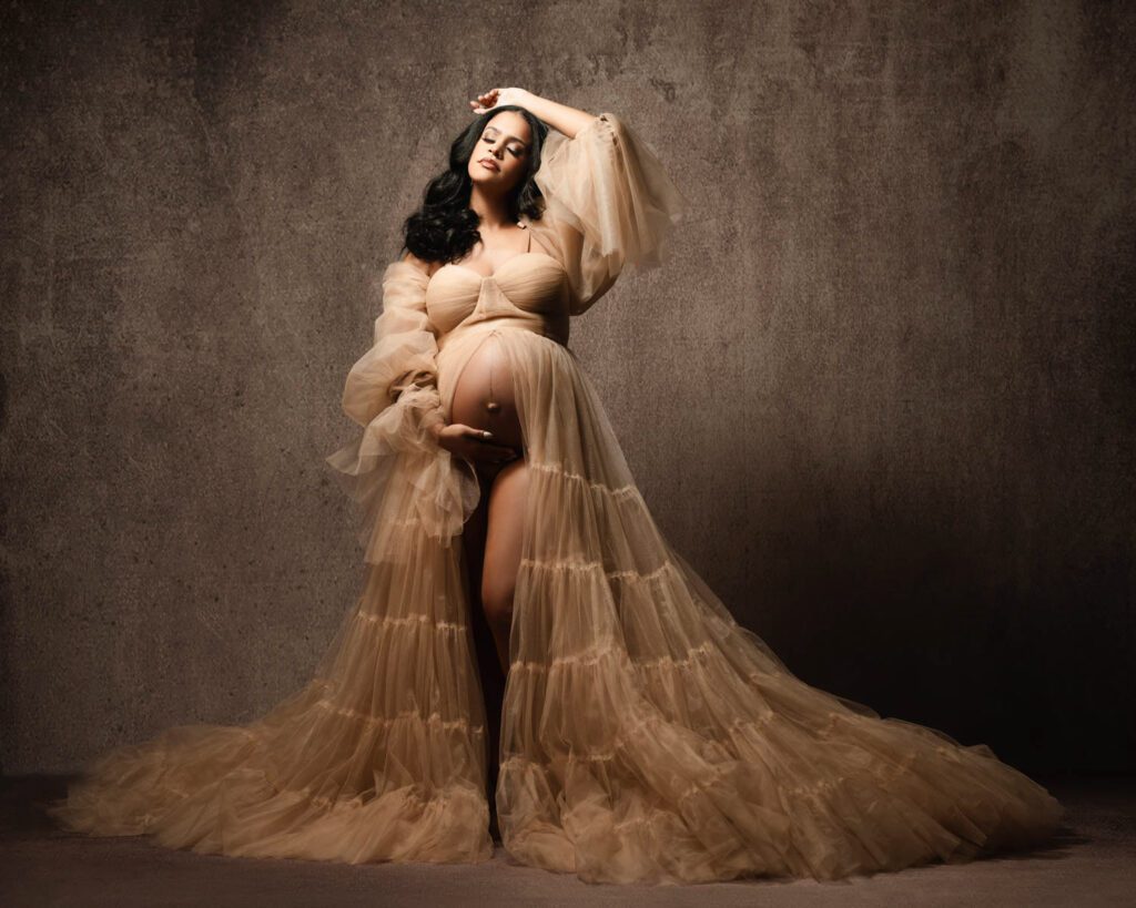 Expecting mother in a dramatic beige tulle gown with one arm raised and the other cradling her bare belly.