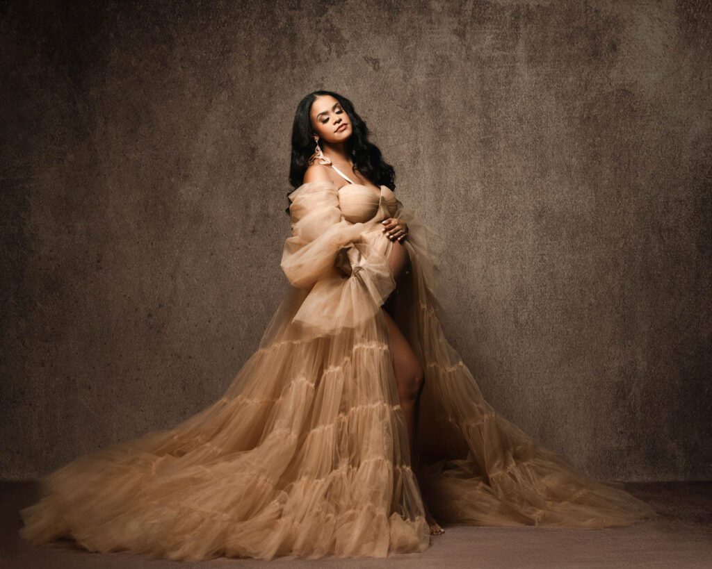 Pregnant woman in a flowing beige tulle gown standing with one hand on her belly against a textured brown backdrop.
