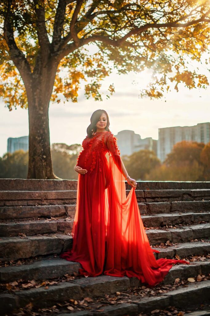 Mom-to-be in a flowing red gown poses on stone steps beneath autumn leaves during a professional maternity session