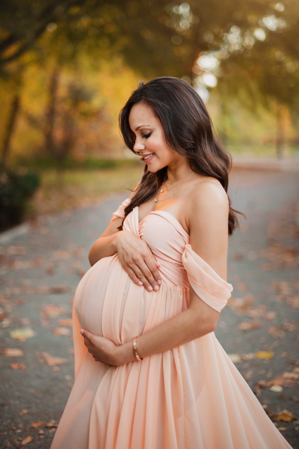 Pregnant woman in a blush off-the-shoulder gown looking down while cradling her belly on an autumn park path at Piedmont Park