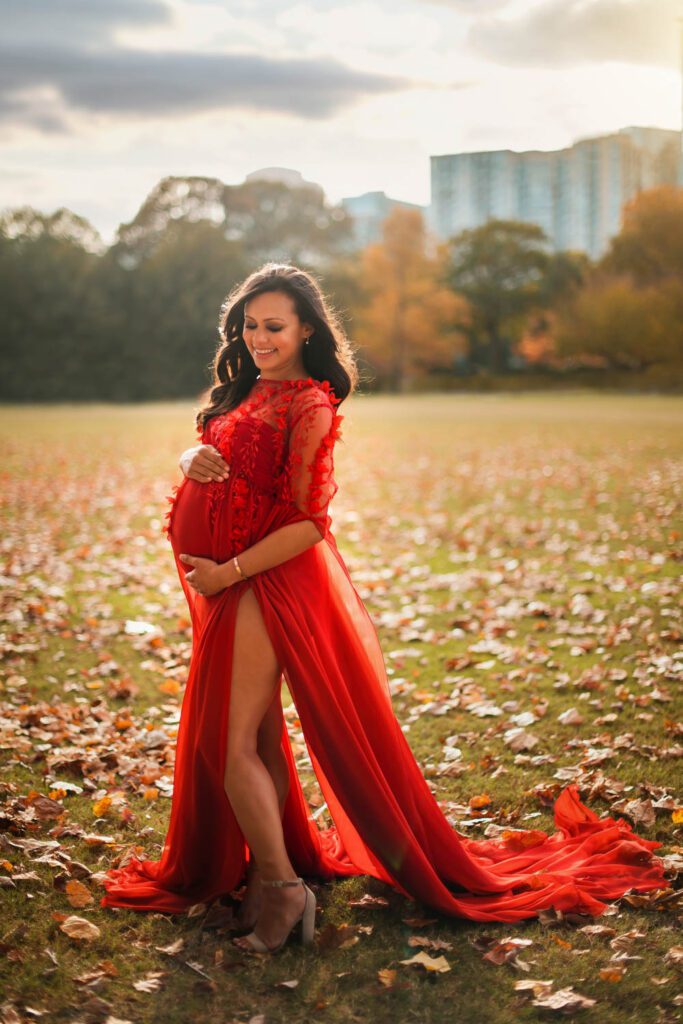 Pregnant woman in a flowing red lace gown standing in The Meadow at Piedmont Park with the Atlanta GA skyline in the background.