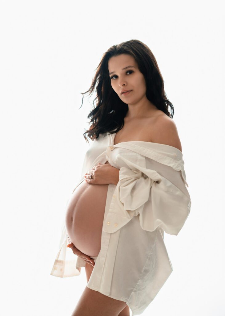Pregnant woman looking at camera in white shirt cradling her belly against a clean white studio background.