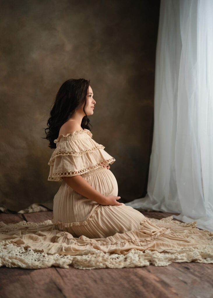 Pregnant mother in cream maternity dress sitting on knees holding baby bump looking out window. 