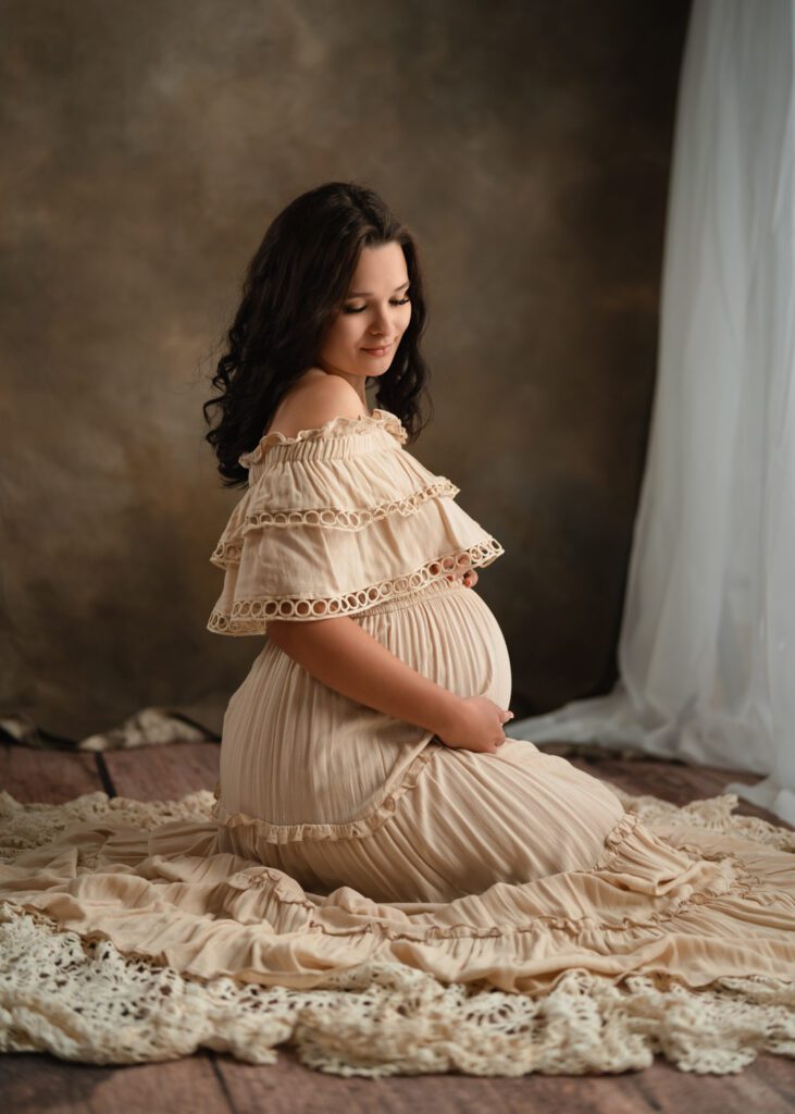 Pregnant woman sitting down on floor in a cream ruffled gown sitting on a textured blanket during a Roswell maternity photo session.
