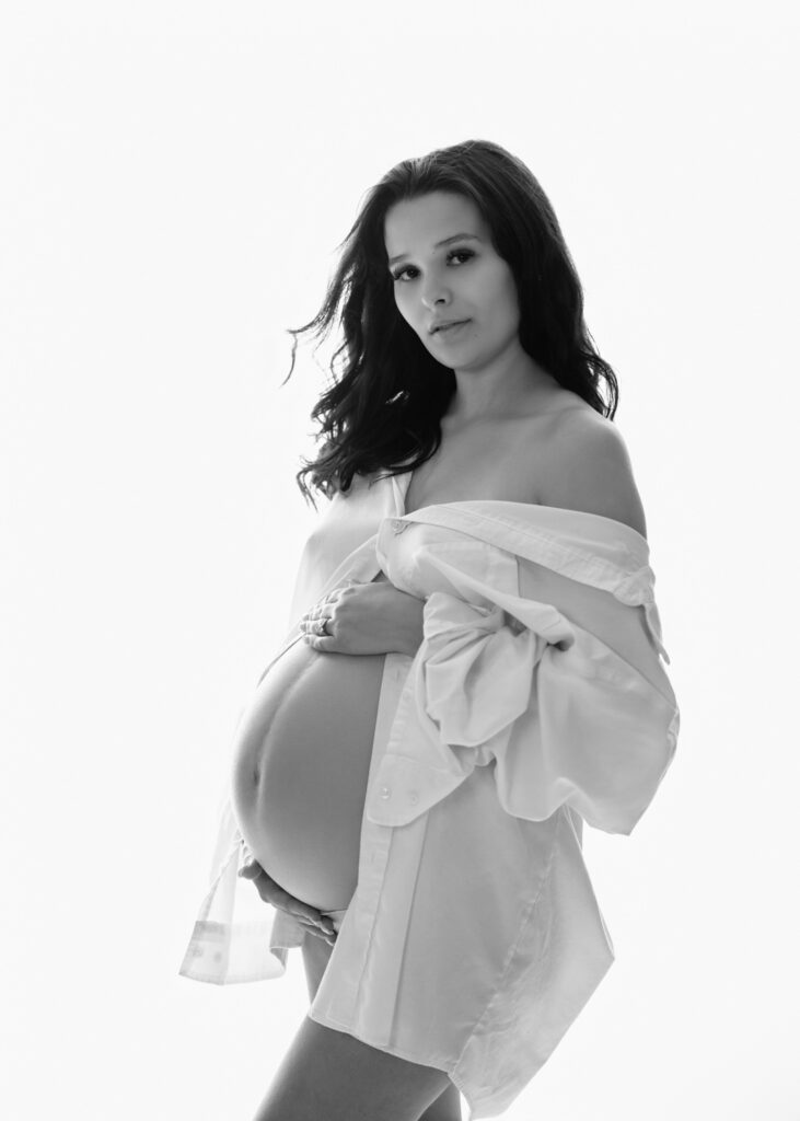 Black and white maternity portrait of an expecting mother in an oversized white shirt cradling her bare belly.