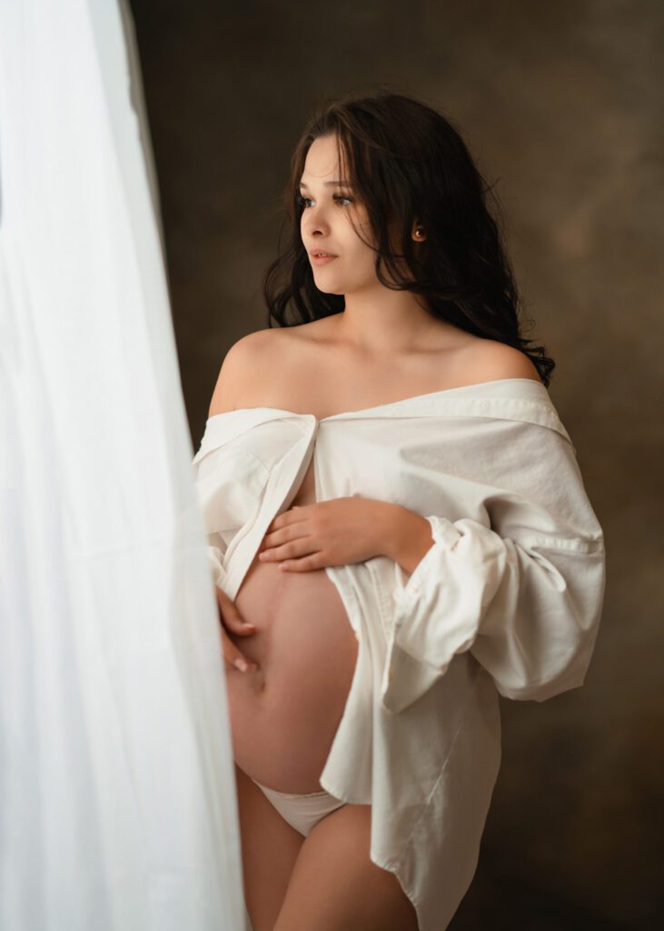 Expecting mother looking out curtain of softly lit window during a Roswell maternity photography session.