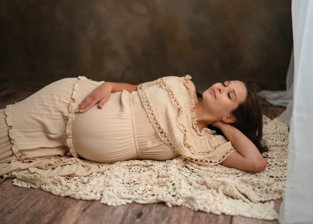 Pregnant woman reclining on a textured studio blanket in a cream gown against a warm brown backdrop.