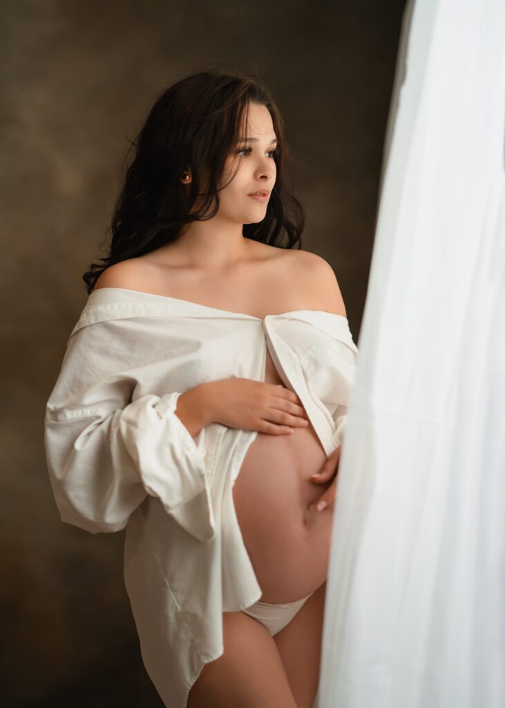 Pregnant mom wearing an oversized white button-up shirt looking out the window photographed by Roswell Maternity Photographer .