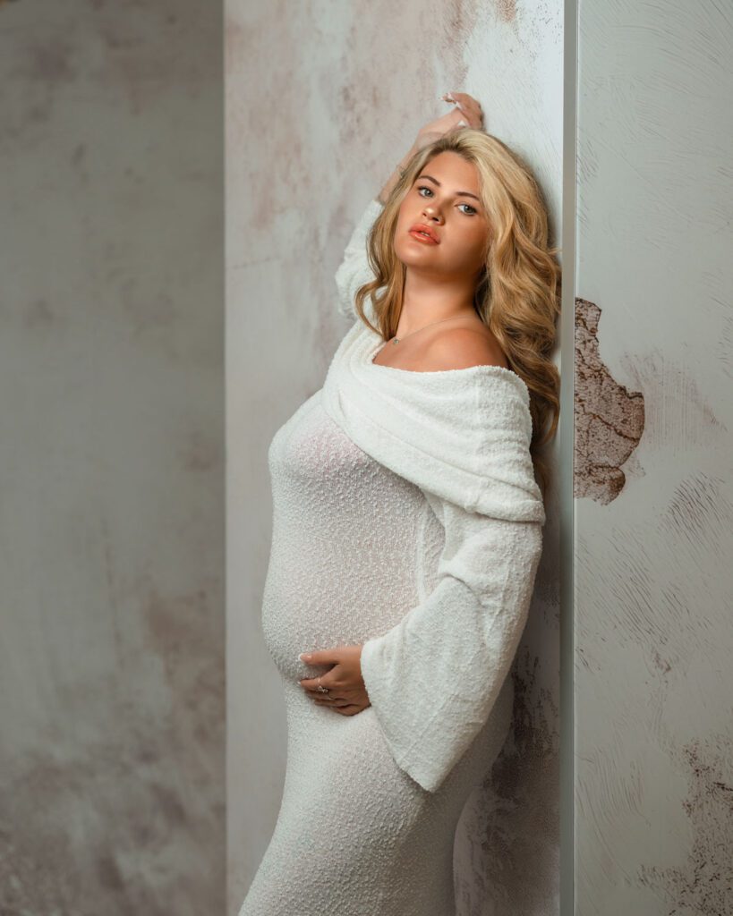 Pregnant woman in a white off-the-shoulder maternity gown posing against a textured studio wall.