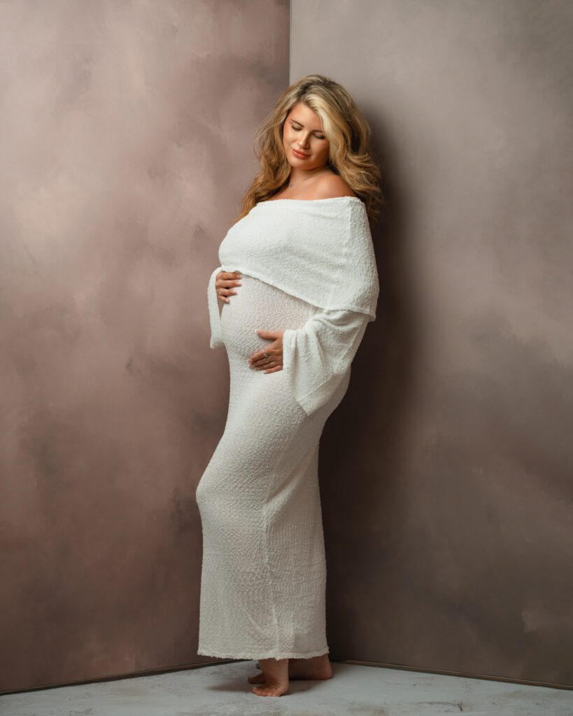 Pregnant woman in a long white dress looking down at her belly, barefoot against a dark textured studio backdrop.