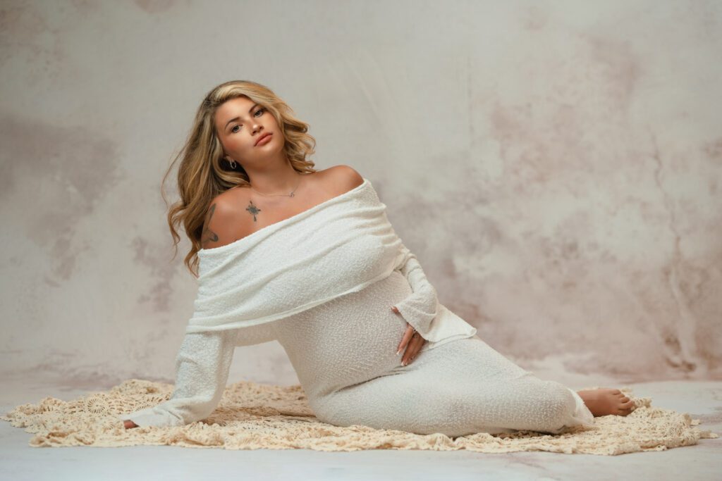 Expecting mom in an off-the-shoulder white gown reclining on a cream crochet blanket while cradling her belly in studio.