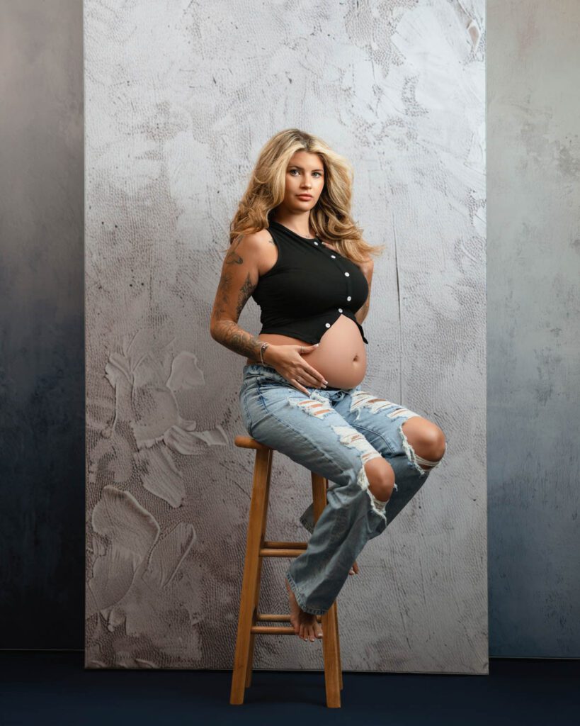 Pregnancy photoshoot inside photo studio with mom sitting on stool wearing ripped blue jeans and black shirt.