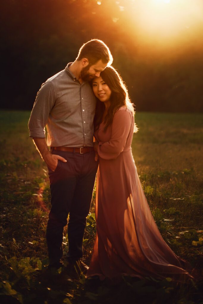 Mom and dad standing close together in dramatic golden sunset light with an open meadow behind them.