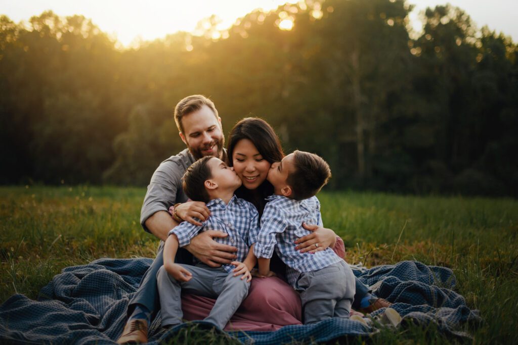 Two sons kissing mom on the cheeks while the family cuddles together on a blanket in the grass.