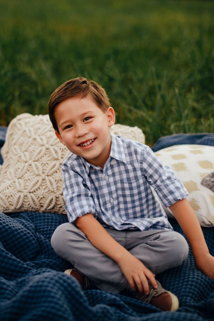 Young boy sitting cross-legged on a blue blanket, smiling with tall grass surrounding him.