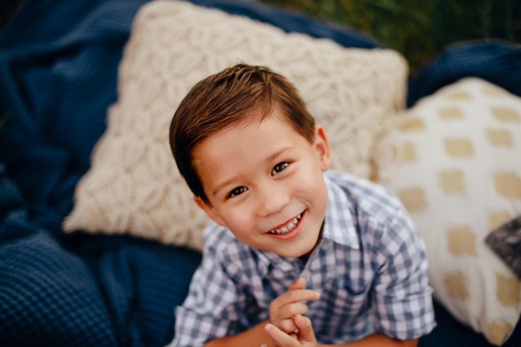 smiling young boy in plaid shirt sitting on blue blanket with textured pillows.