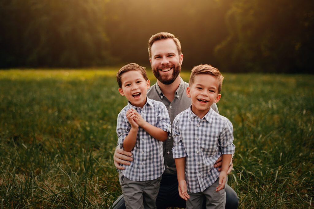 dad and his twin sons kneeling down in grass at sunset with pond and stone wall in the background.