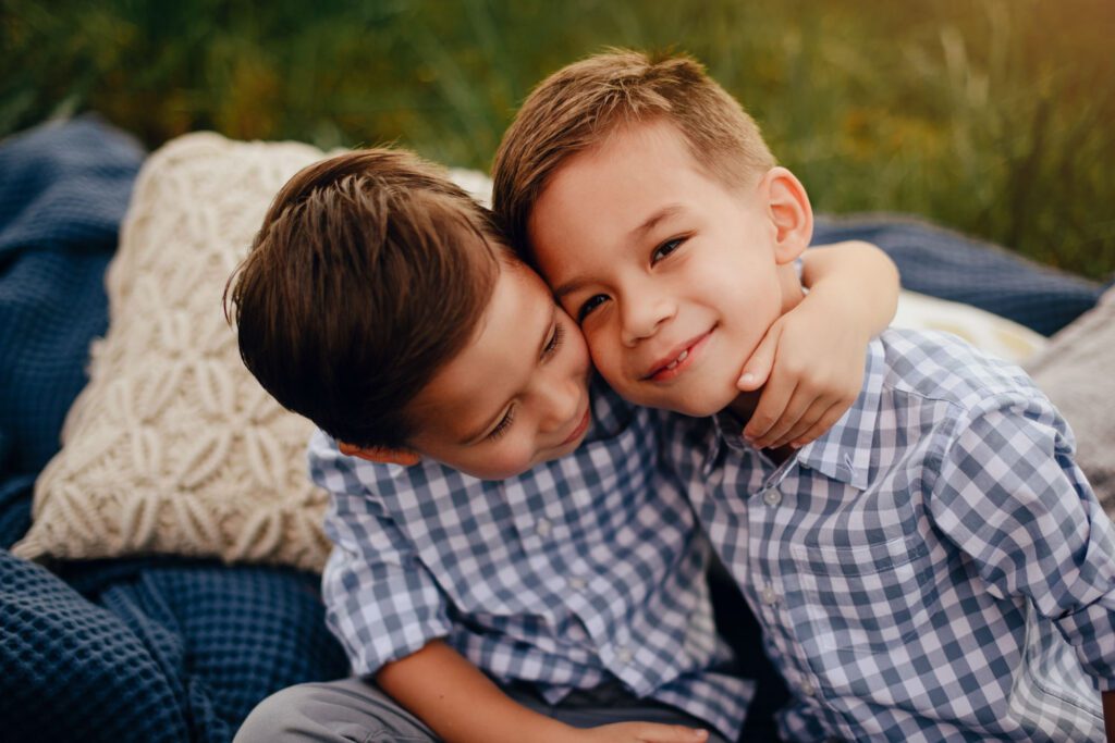 brothers hugging in plaid shirts and jeans seated on blue textured blanket on grass