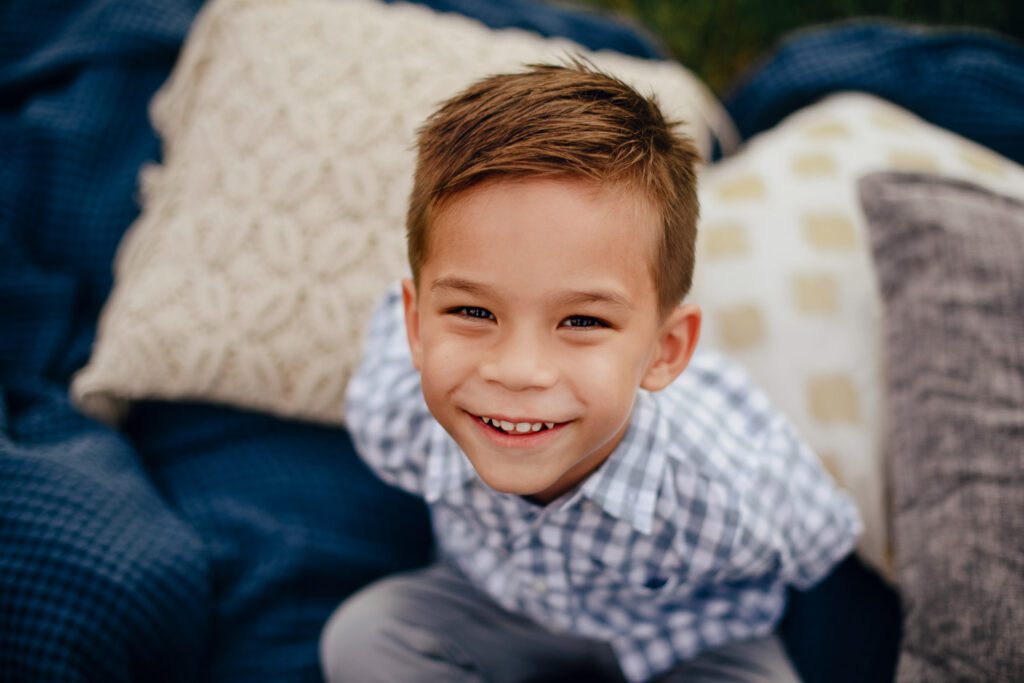 smiling young boy in jeans sitting on blue blanket with cream pattern pillows.