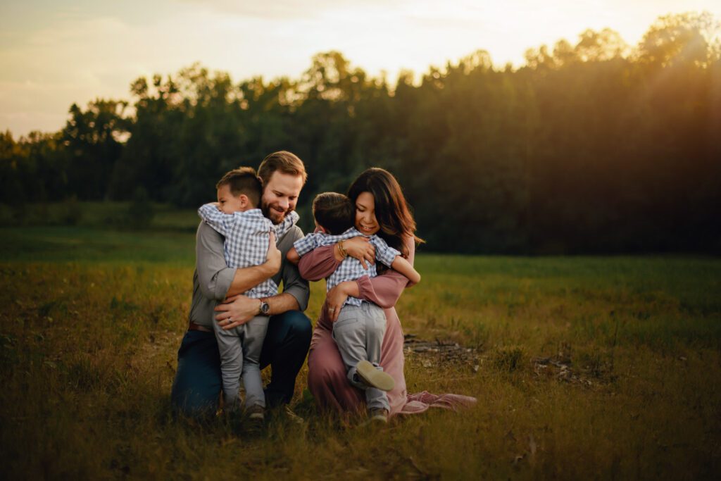 Parents kneeling in the meadow while hugging their two sons in warm golden evening light.