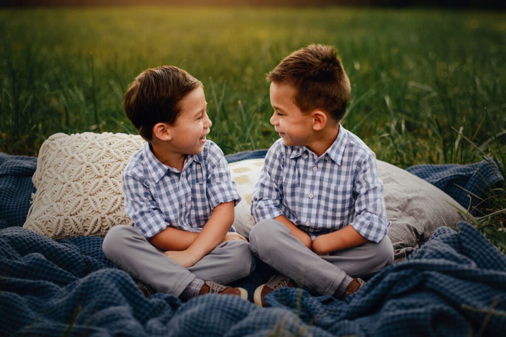 Brothers sitting cross-legged on a blanket facing one another and smiling in the grassy meadow.