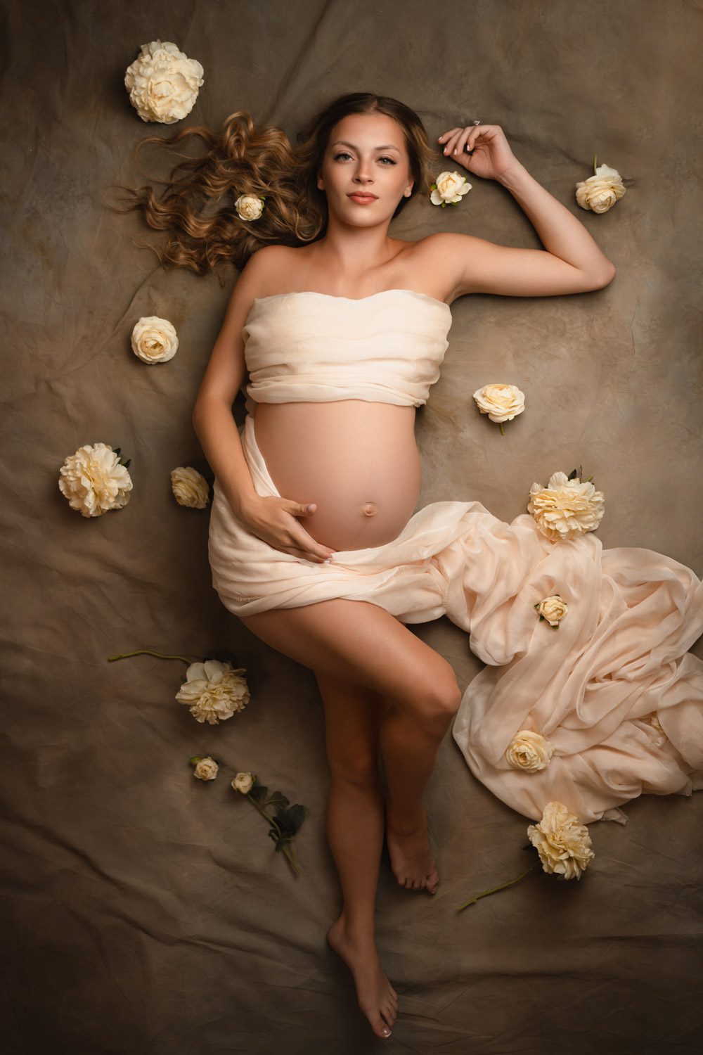 Overhead maternity portrait of an expecting mother reclining among soft fabric and cream flowers.