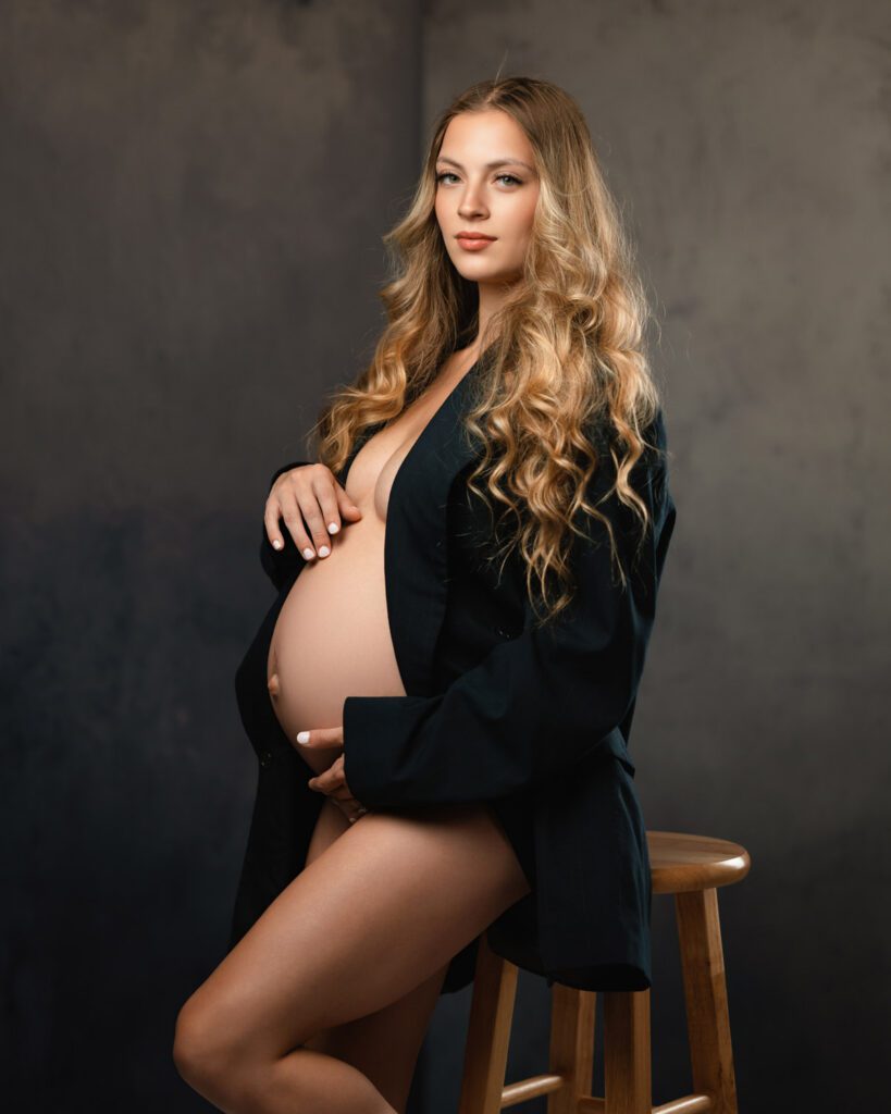 Mom-to-be seated in an open black blazer with one hand resting above her belly.