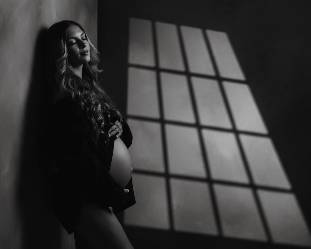 Black and white maternity portrait in an open black blazer with windowpane shadows across the studio wall.