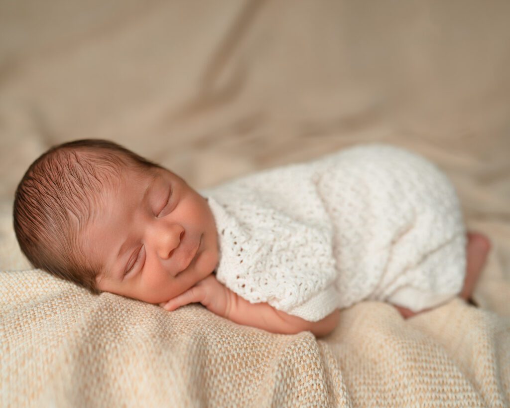 Sleeping newborn resting on his tummy in a cream crochet romper on a textured tan blanket.