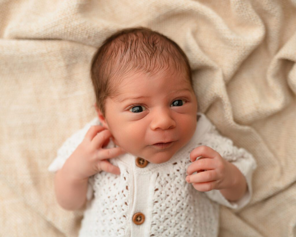 Close-up of a newborn baby in a cream knit romper with brown buttons, lying on his back with wide eyes.