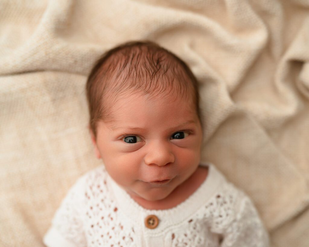 Newborn baby looking toward the camera with bright eyes while resting on a soft neutral blanket.