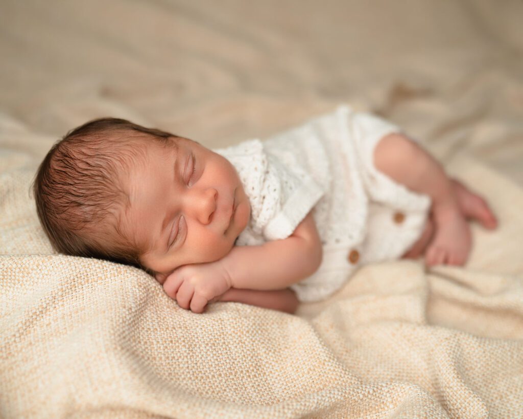 Sleeping infant with hands tucked under his cheek on a textured tan blanket for Alpharetta newborn photos.