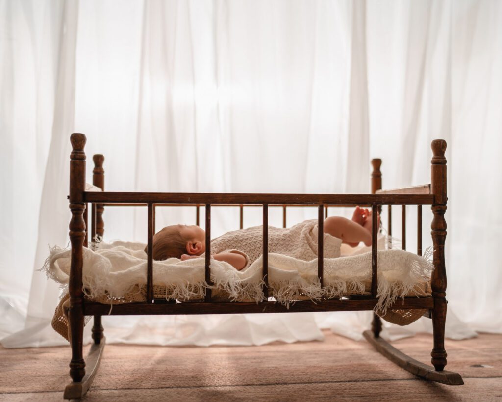 Newborn baby lying on his back in an antique wooden cradle with soft window light and cream blankets.