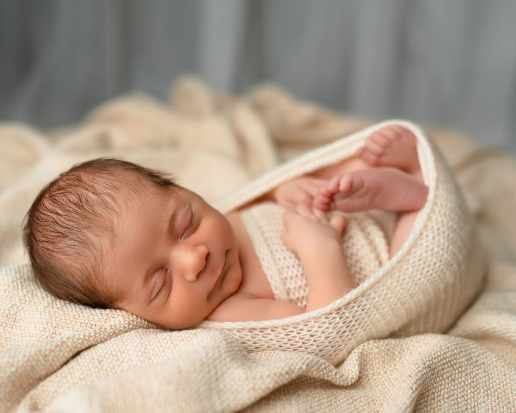 Newborn baby bundled in a cream knit swaddle with one foot peeking out while resting in a wooden bowl.