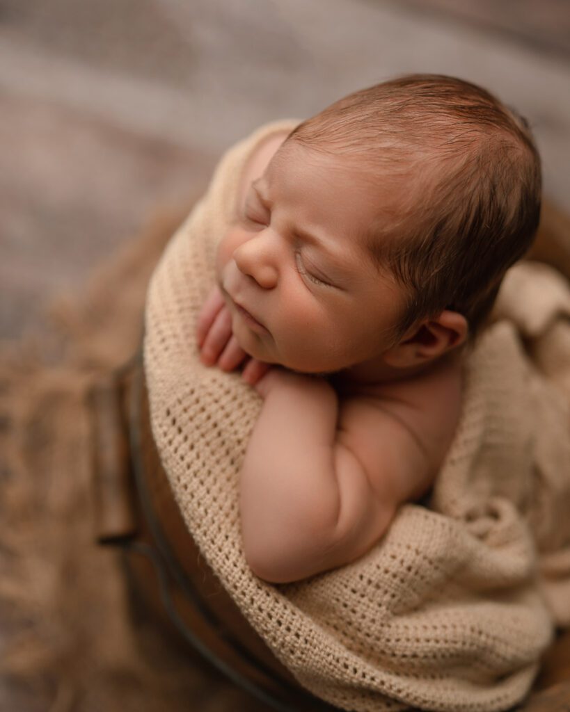 Sleeping newborn resting in a wooden bucket with his head on his hands and a cream knit wrap.