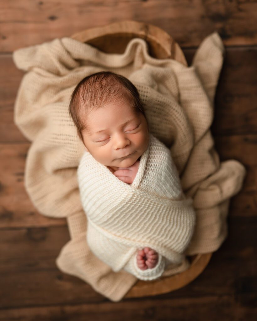 Newborn baby wrapped in a neutral knit swaddle and resting in a wooden bowl for a newborn photographer in Alpharetta.