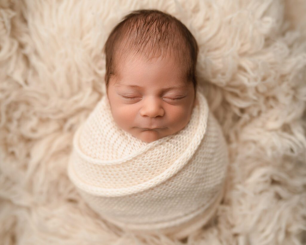 Sleeping newborn bundled in a neutral knit wrap and posed on a soft cream flokati blanket.