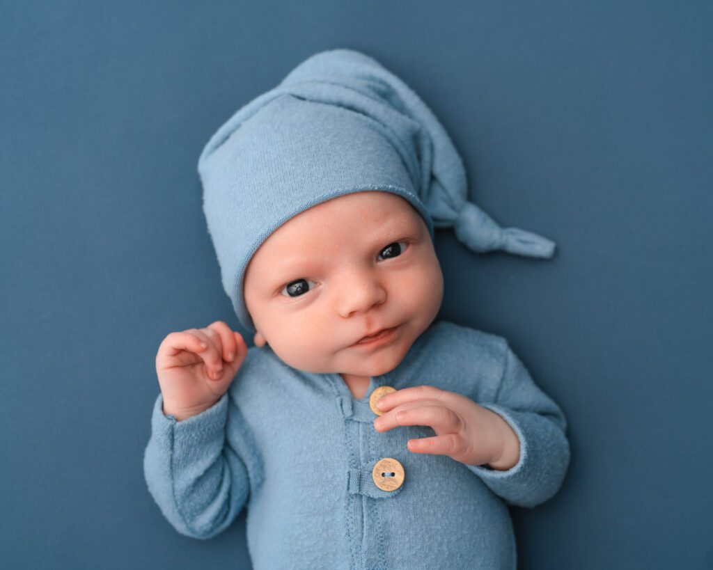 Alert newborn baby boy looking at the camera in a light blue outfit against a matching blue backdrop.