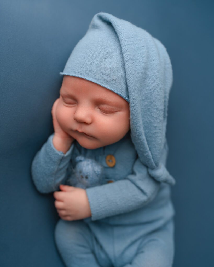 Sleeping newborn baby boy in a soft blue romper and sleepy cap, resting his head on one hand.