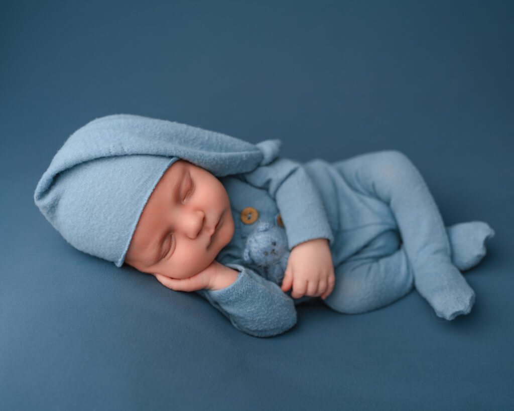 Sleeping baby boy in a light blue romper cuddling a blue knit teddy bear on a soft blue backdrop.