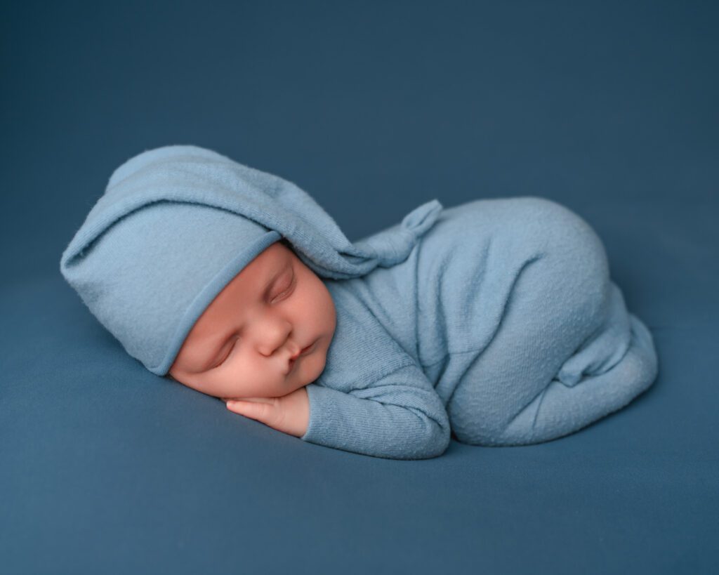 Sleeping newborn baby boy in a light blue footed romper and sleepy cap posed on a matching blue backdrop.